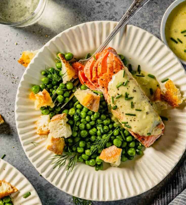 white scalloped plates filled with green peas, salmon topped with butter, croutons and herbs. Glasses of wine set around