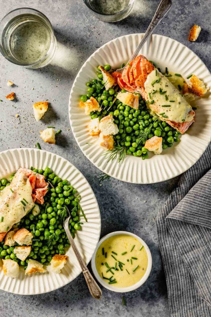 white scalloped plates filled with green peas, salmon topped with butter, croutons and herbs. Glasses of wine set around