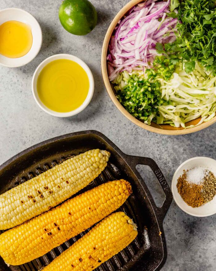 a bowl filled with shredded green cabbage, sliced red onion, cilantro and jalapeno. Corn charred in a grill pan and honey, olive oil and lime set off to the side