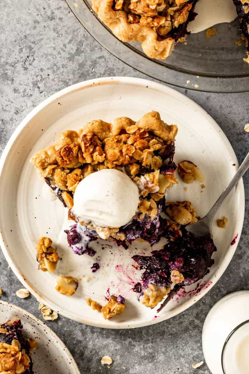 a slice of blueberry crumble pie on a white plate with a scoop of ice cream on top