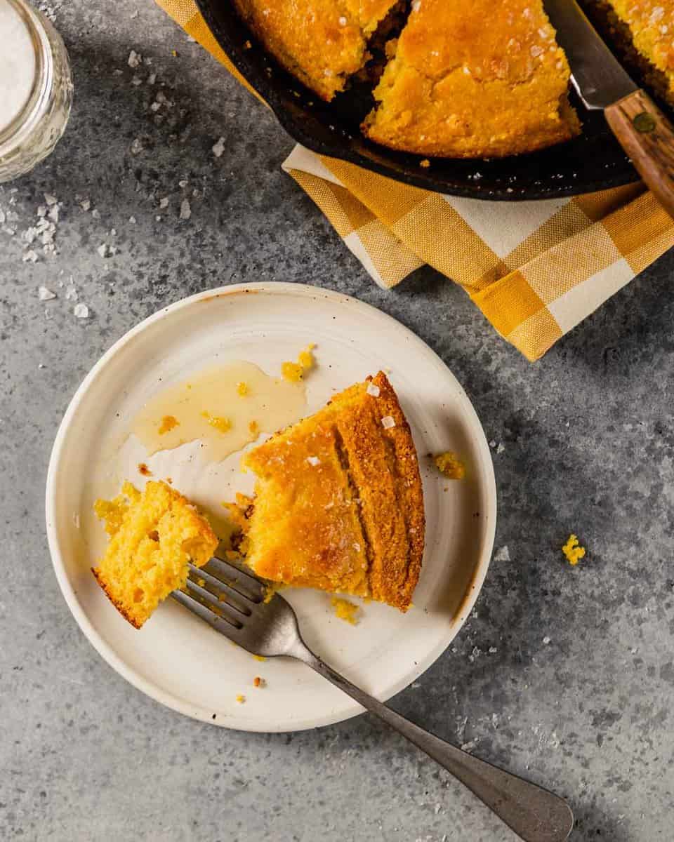 a piece of cornbread on a white place with a skillet of cornbread off to the side