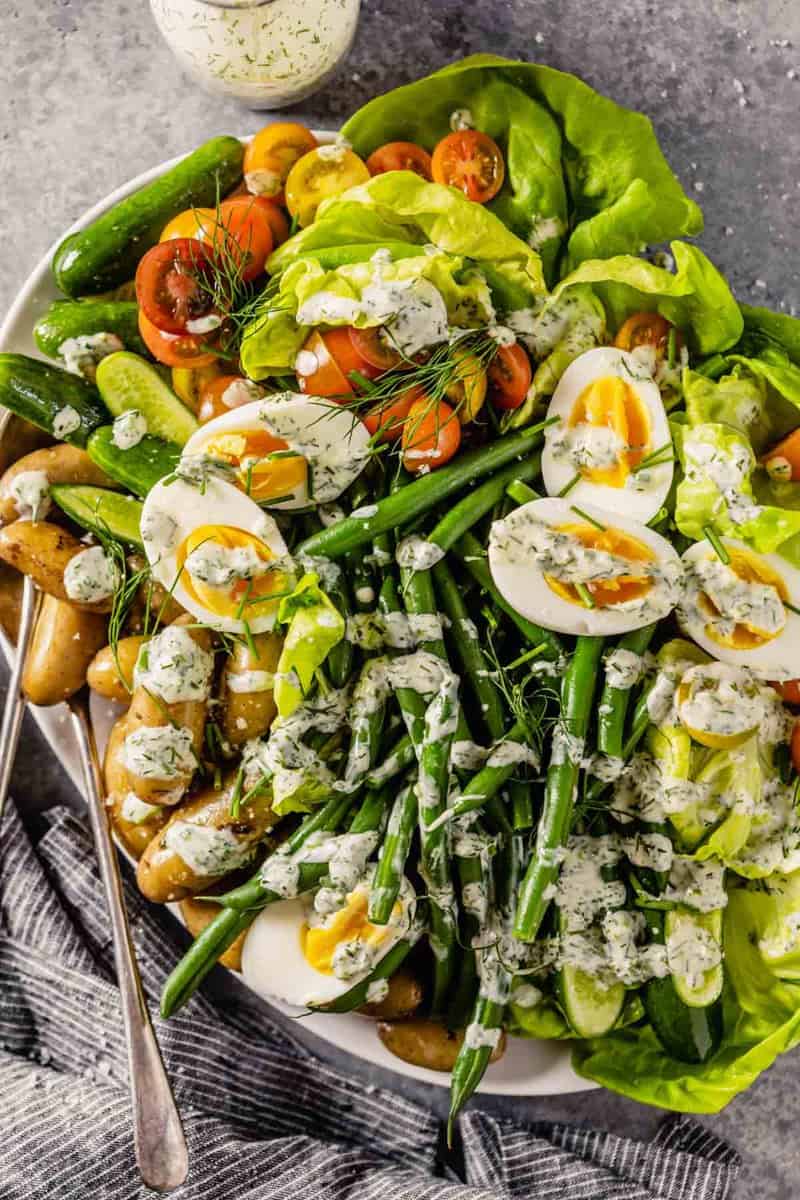 large white platter filled with butter lettuce, blanched green beans, fingerling potatoes, halved cherry tomatoes, mini cucumbers, hard boiled eggs and a creamy ranch dressing