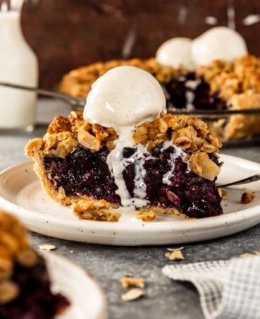 piece of blueberry pie on a white plate with a scoop of melty vanilla ice cream on top