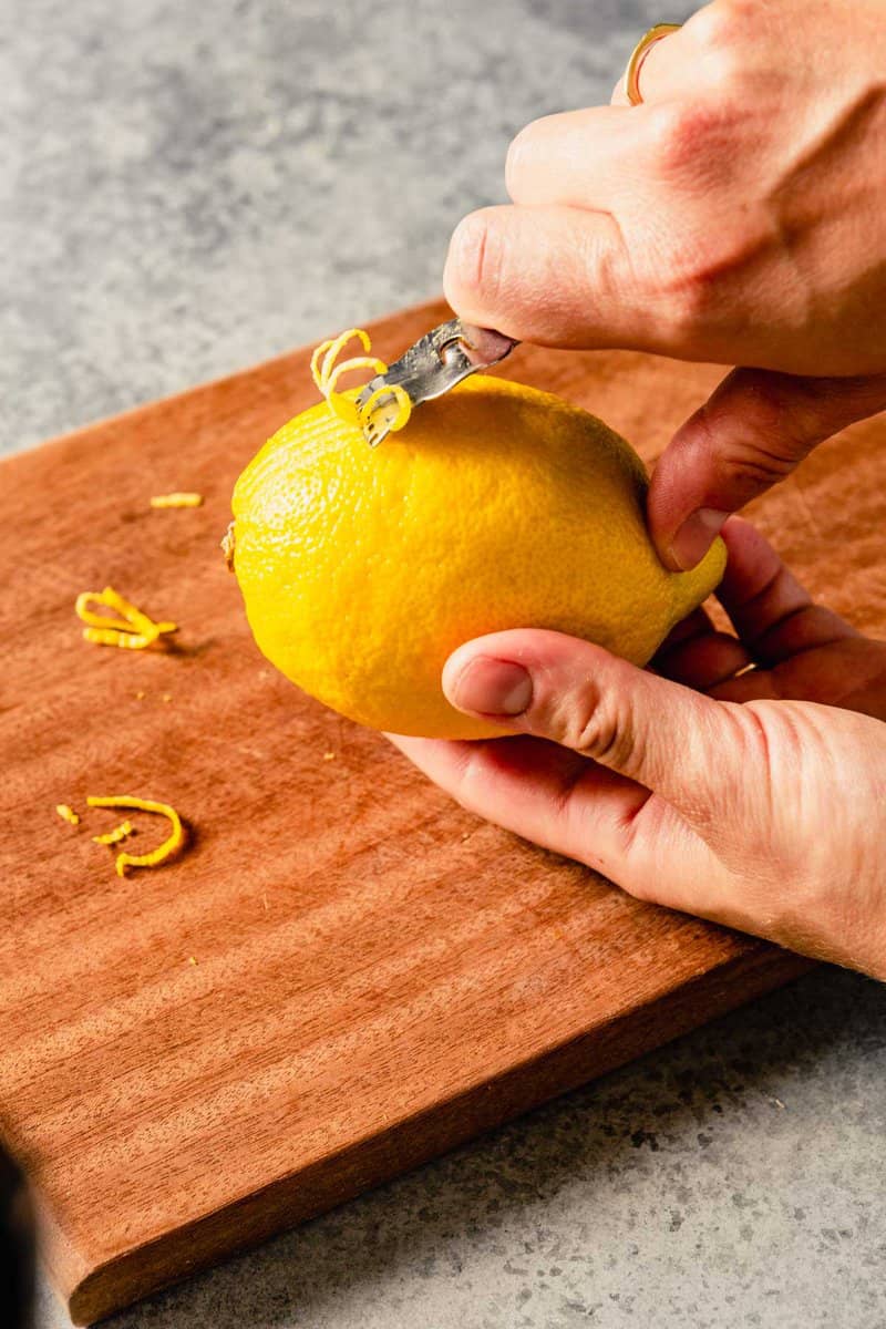 two hands zesting a lemon using a citrus zester set over a wood cutting board