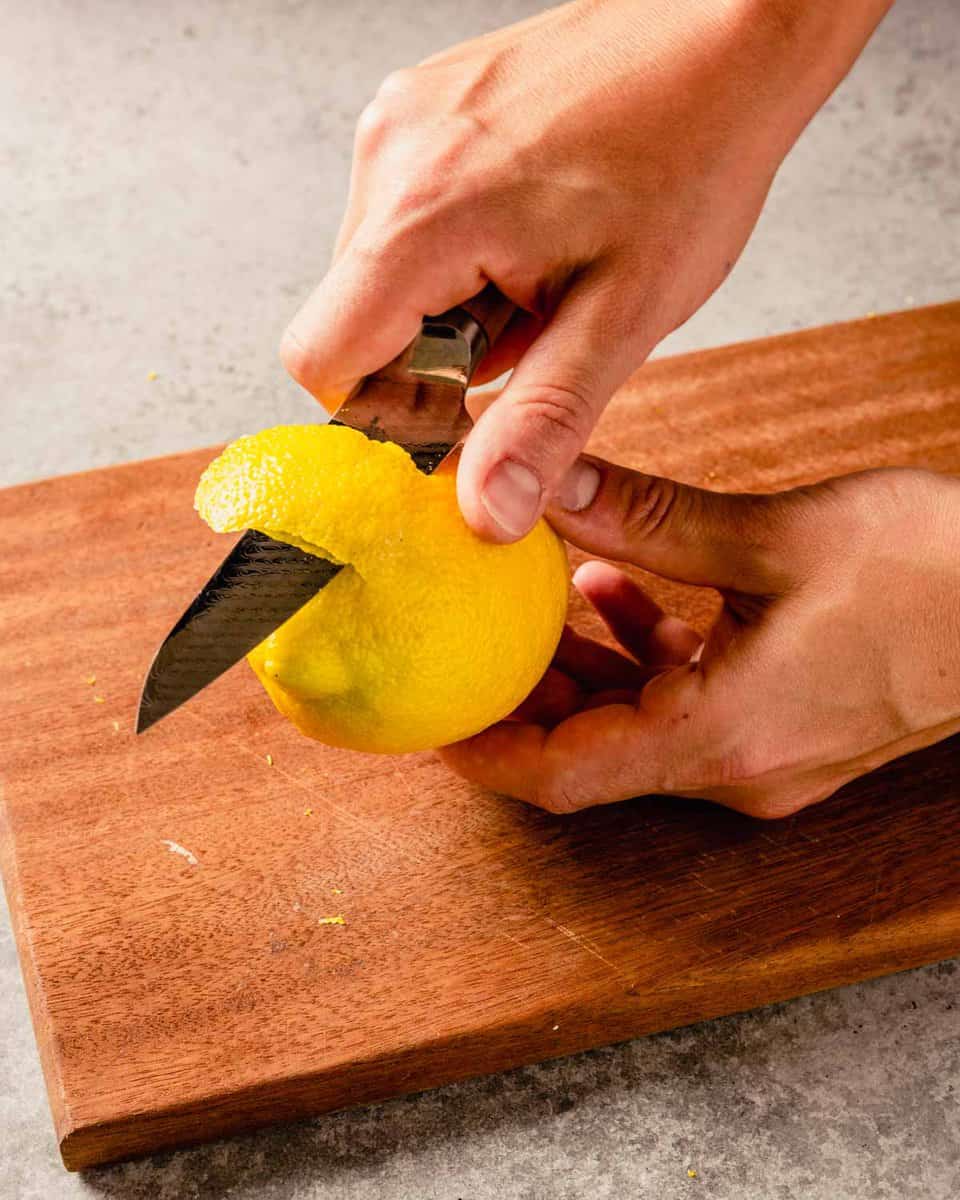 two hands peeling lemon zest from a lemon with a paring knife