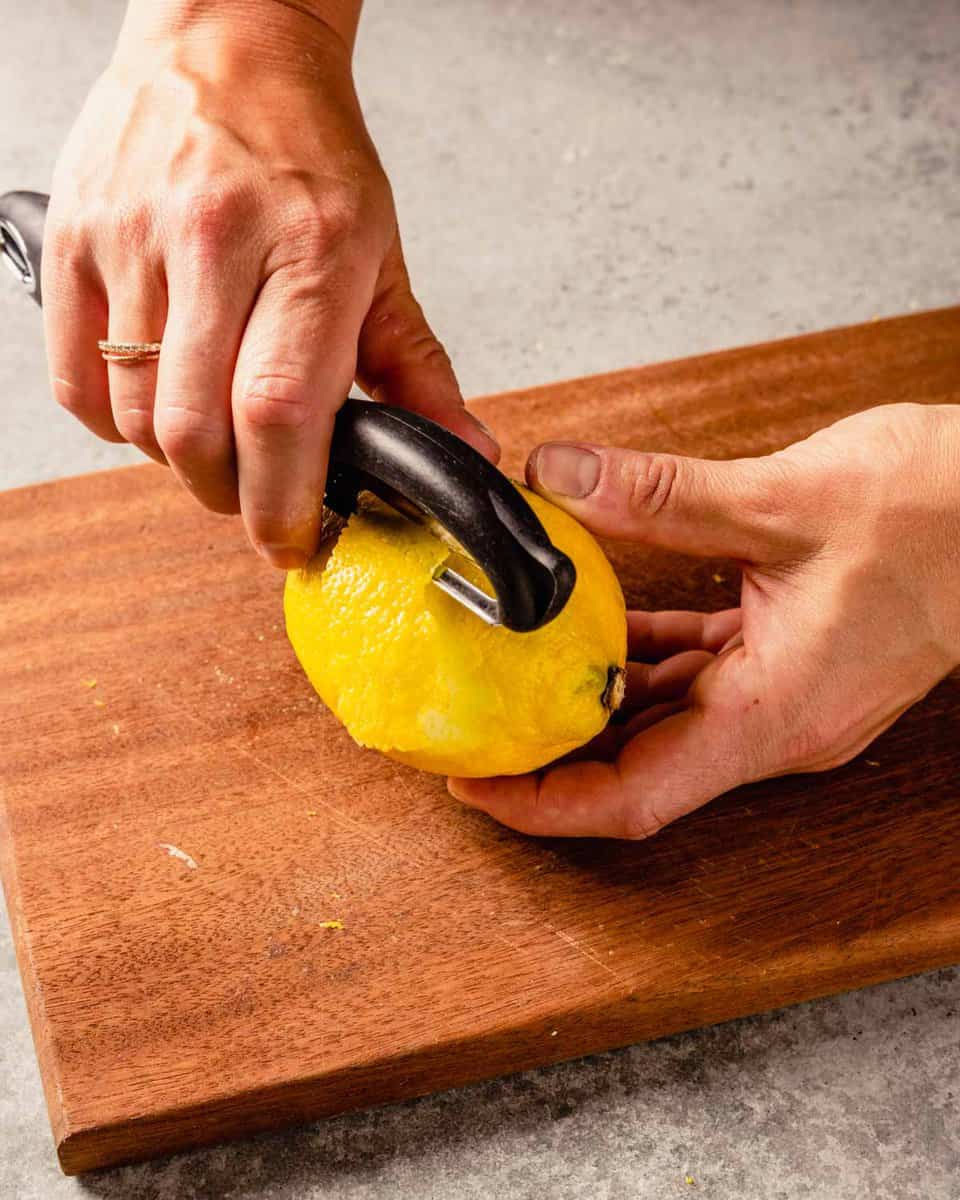 two hands peeling lemon zest from a lemon with a vegetable peeler