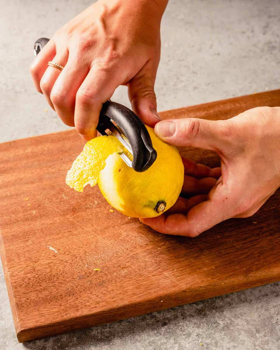 two hands peeling lemon zest from a lemon with a vegetable peeler