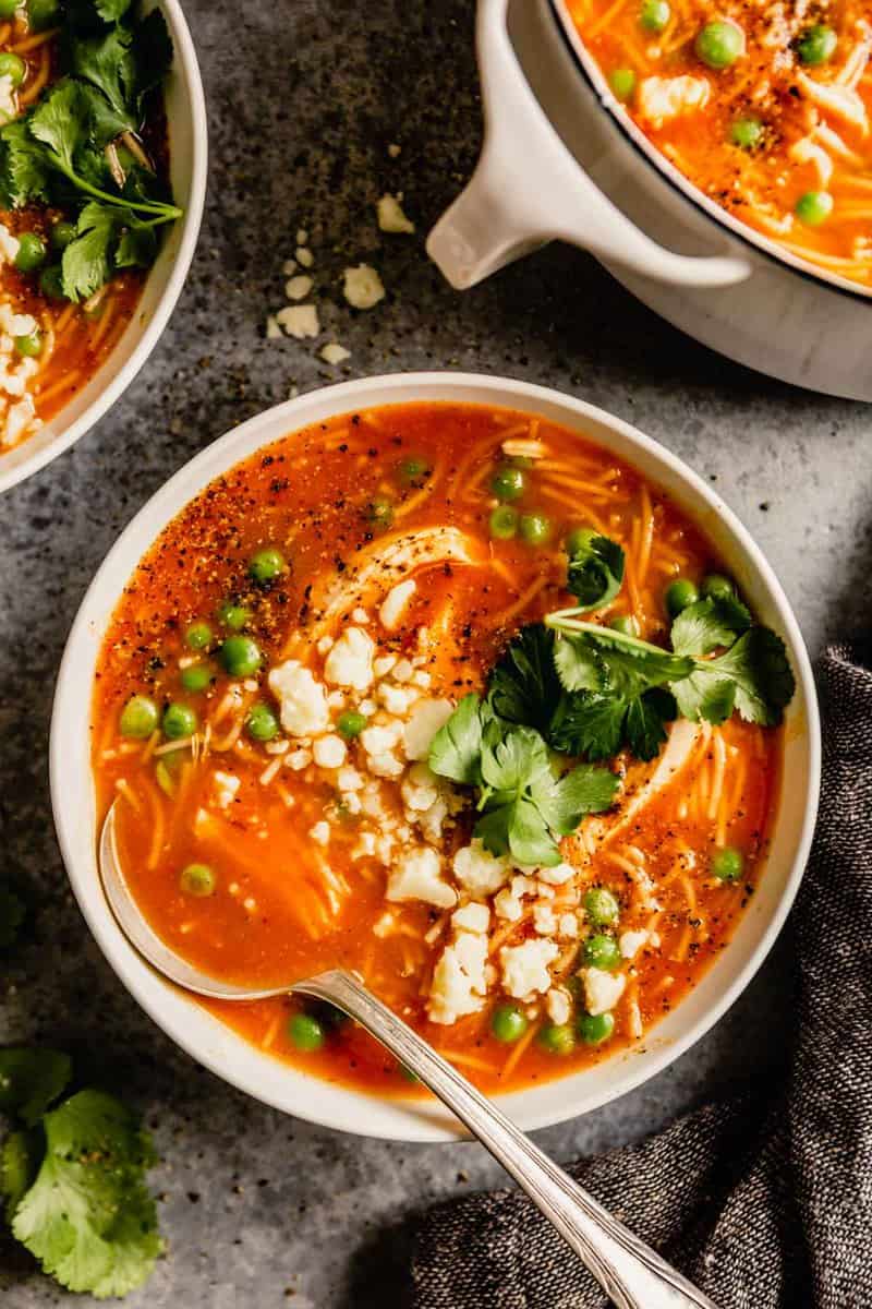 tomato soup with shredded chicken, peas, cilantro and crumbled cheese set on a table with a white pot filled with soup