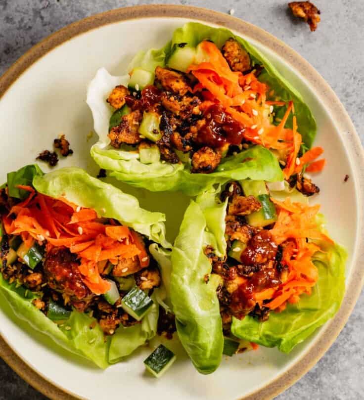 three lettuce wraps on a white plate. lettuce wraps toped with shredded carrots and chili sauce