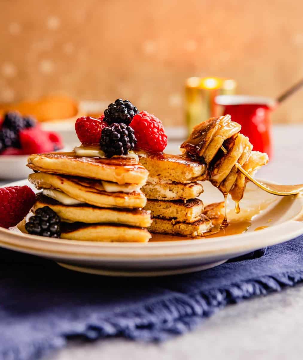 whole wheat pancakes stacked on a plate with berries on top and maple syrup dripping down