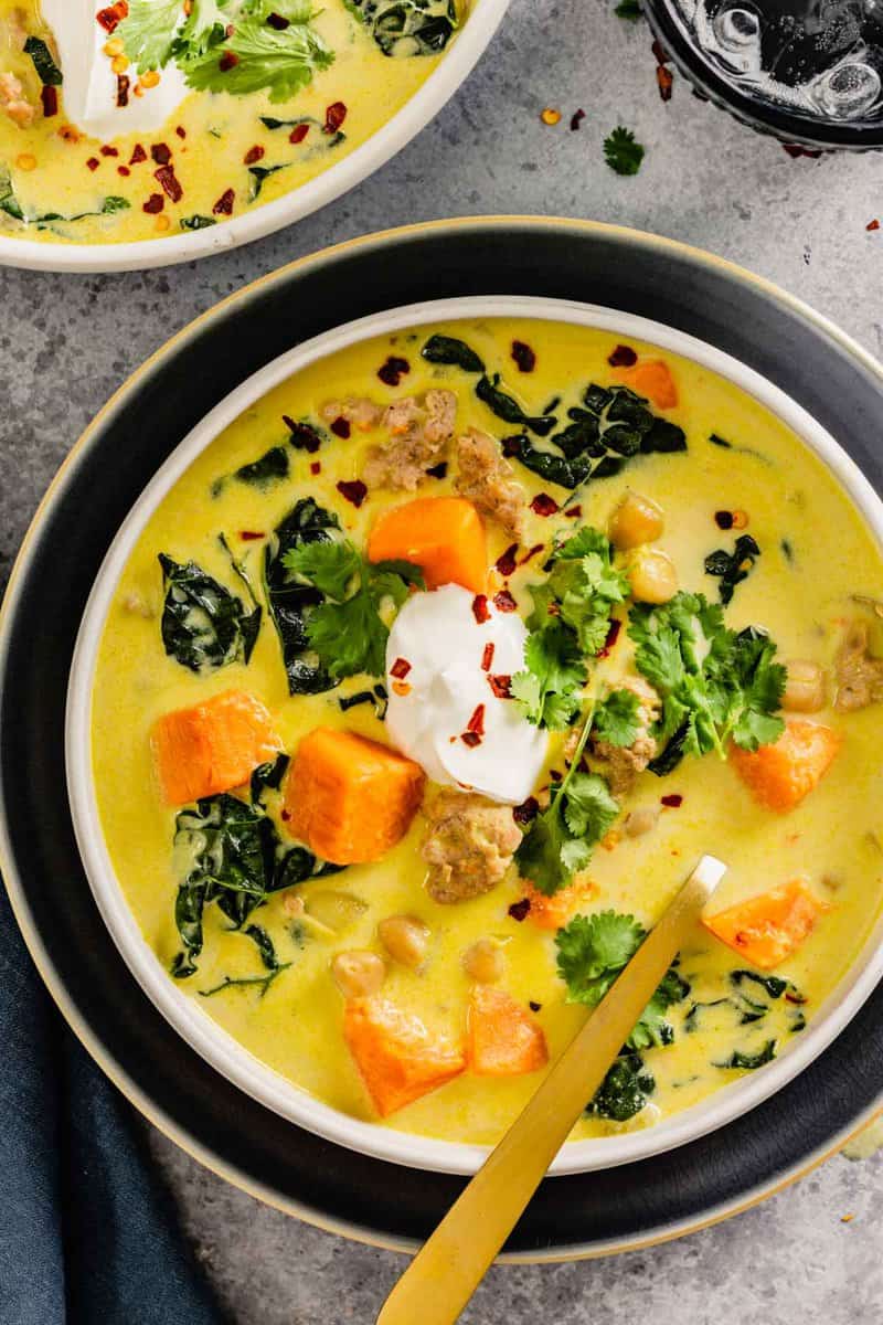 shallow white bowl set on a dark blue plate filled with a yellow-colored curry soup with chunks of sweet potato, kale, ground pork and chickpeas. topped with cilantro, yogurt, and red pepper flakes.