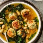 yellow-orange broth in a white bowl with meatballs, kale and beans