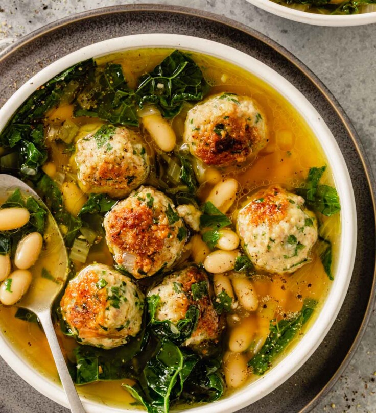 yellow-orange broth in a white bowl with meatballs, kale and beans
