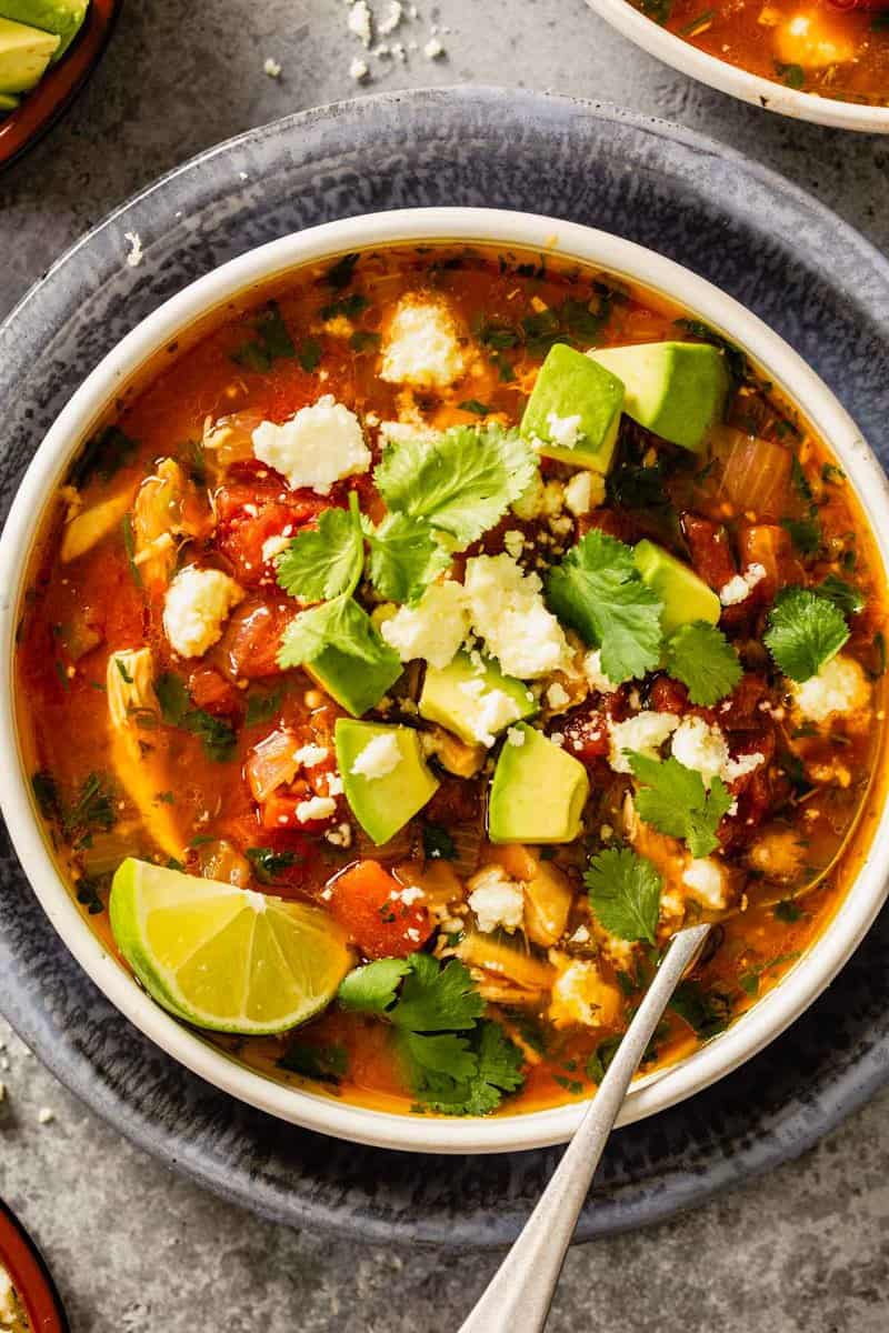 brothy soup in a white bowl with shredded chicken, tomatoes, diced avocado, crumbled cheese and cilantro