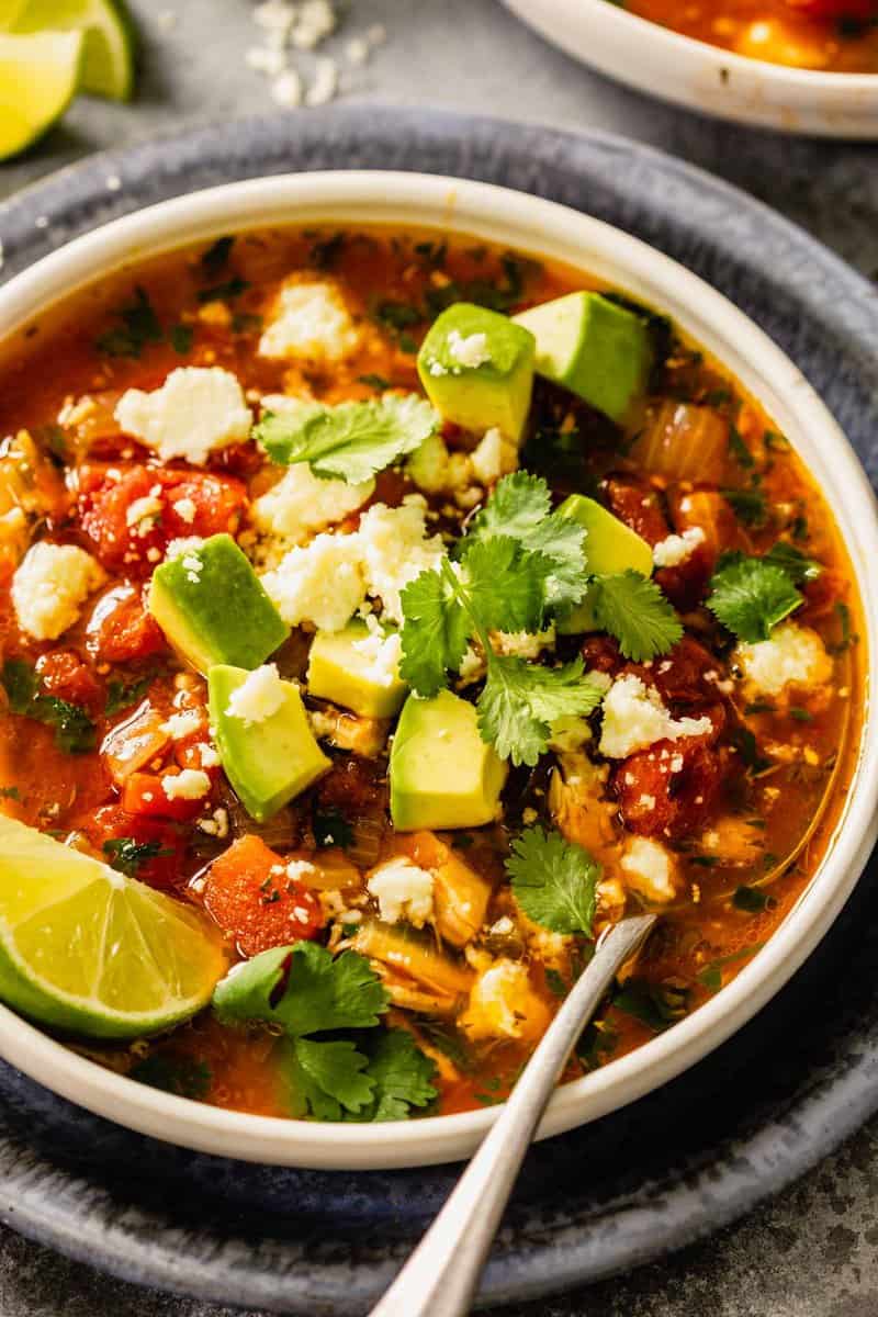 brothy soup in a white bowl with shredded chicken, tomatoes, diced avocado, crumbled cheese and cilantro