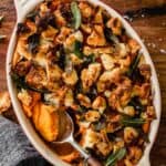 oval casserole dish with a serving spoon set in it with sweet potato casserole topped with croutons and sage