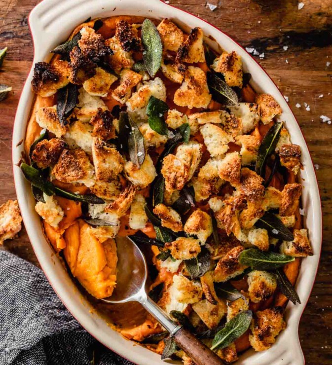 oval casserole dish with a serving spoon set in it with sweet potato casserole topped with croutons and sage