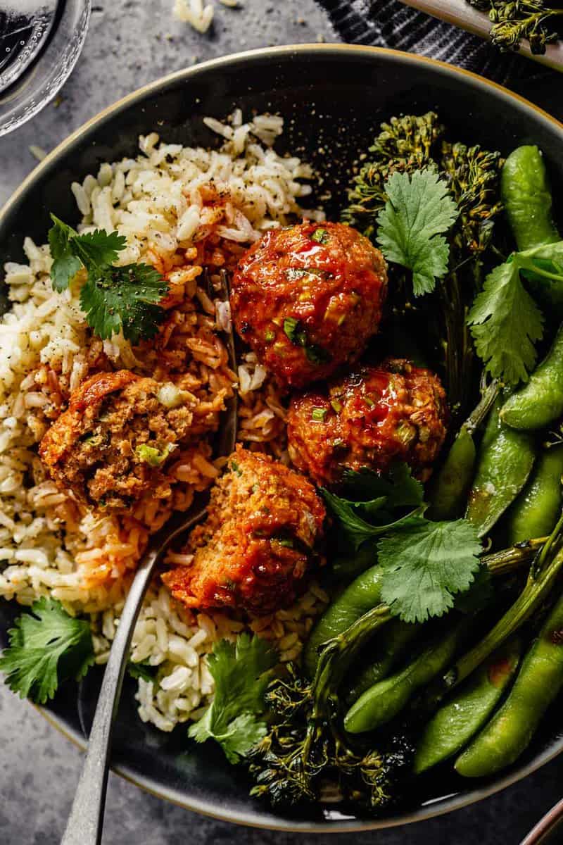 meatballs coated in a red glaze set on top of rice with broccolini and edamame on a blue plate