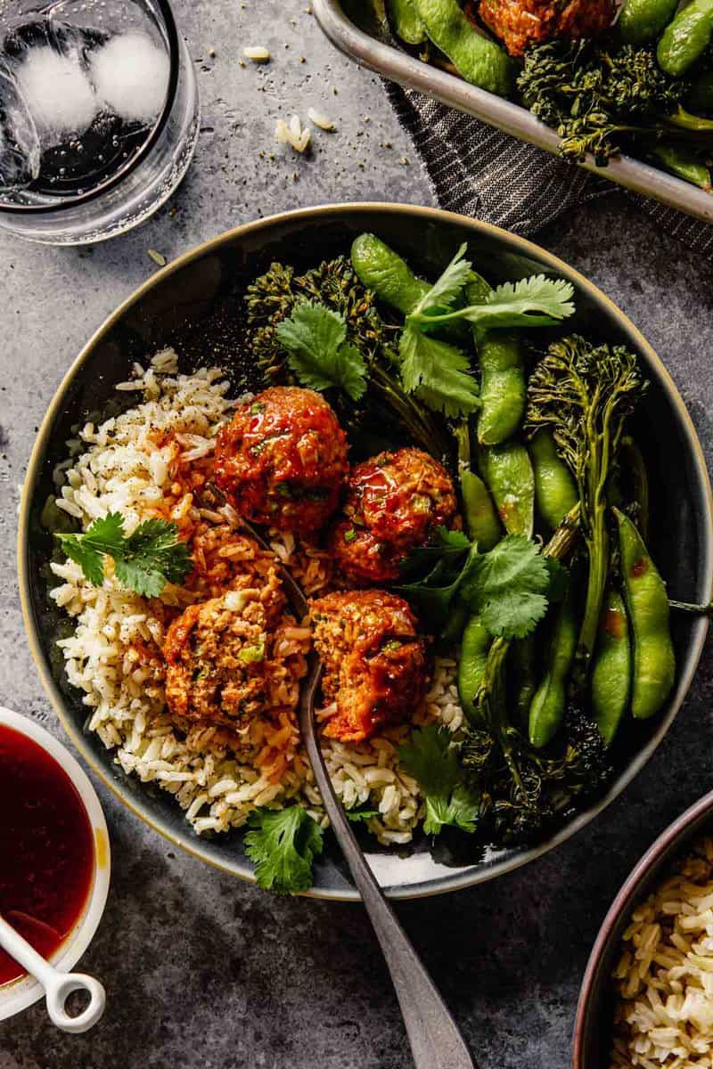 meatballs coated in a red glaze set on top of rice with broccolini and edamame on a blue plate