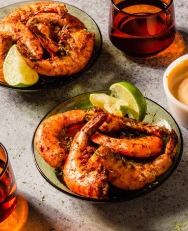 large in-shell shrimp coated in spices and in a green glass serving bowl with orange glassware set around and a dipping sauce in a white bowl