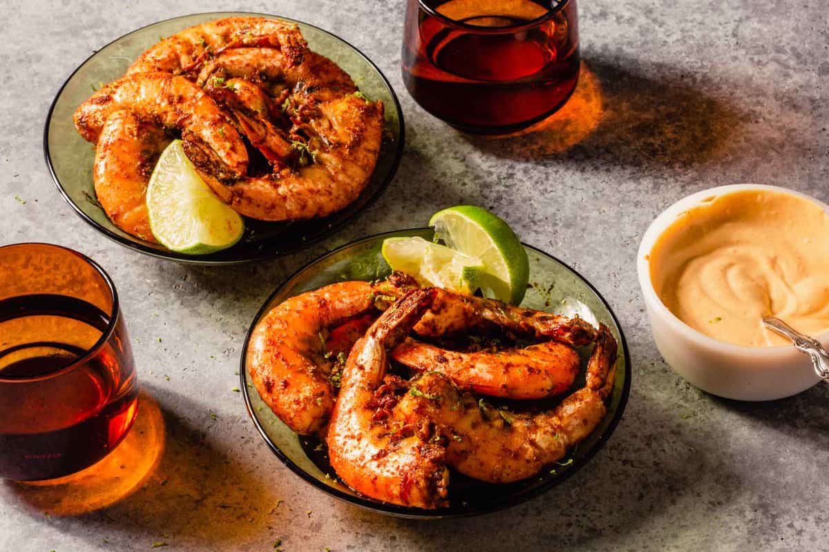 large in-shell shrimp coated in spices and in a green glass serving bowl with orange glassware set around and a dipping sauce in a white bowl