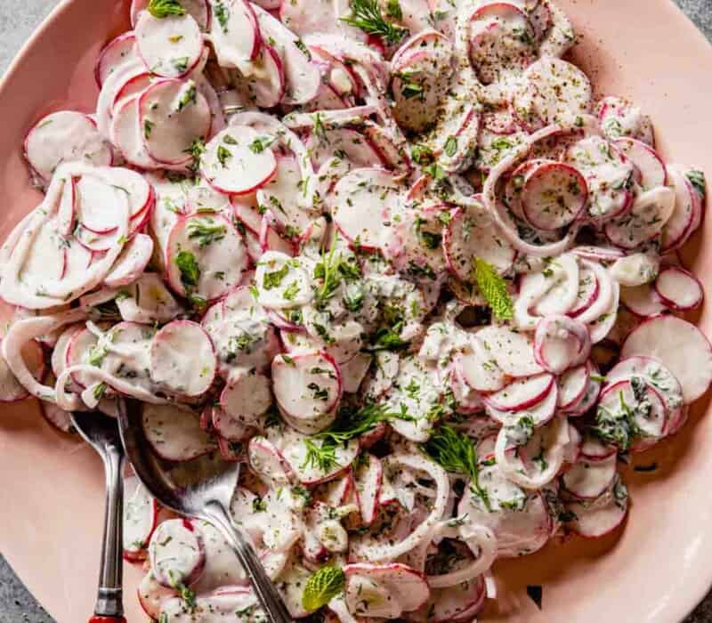 thinly sliced radishes and shallot coated in a creamy herb dressing on a pink plate with two red-handled spoons