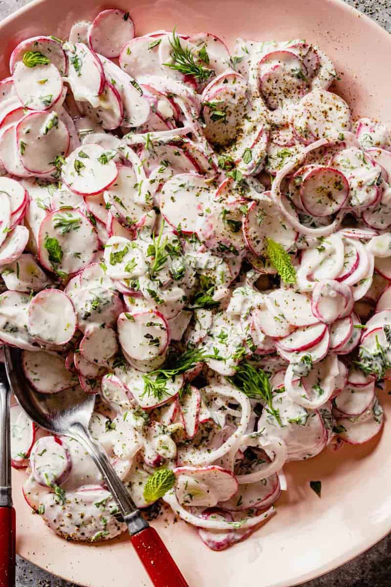 thinly sliced radishes and shallot coated in a creamy herb dressing on a pink plate with two red-handled spoons