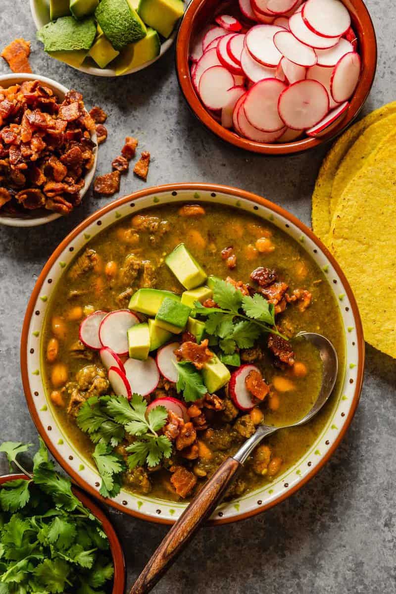 beef and bean stew in a multi-colored bowl topped with avocado, cilantro, radish and bacon bits