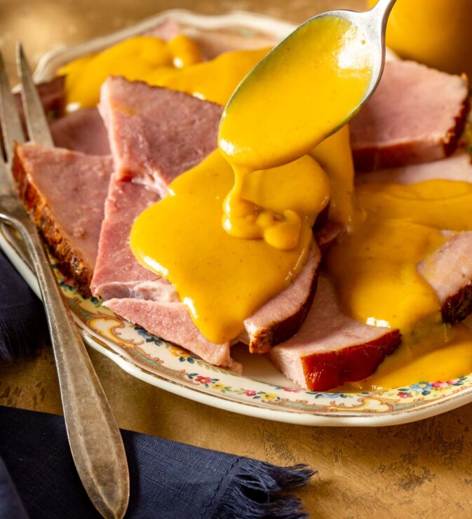 a silver spoon drizzling yellow mustard sauce over slices of ham on an ornate platter