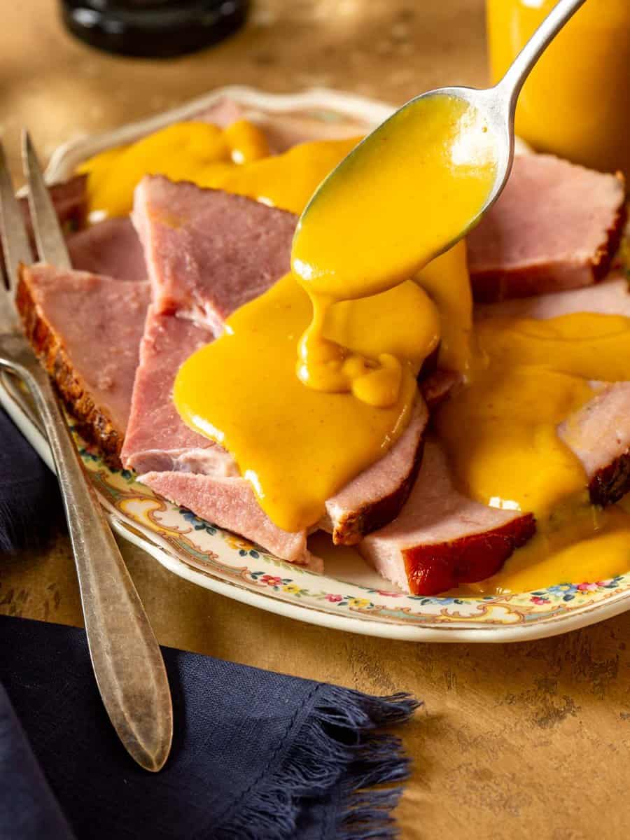 a silver spoon drizzling yellow mustard sauce over slices of ham on an ornate platter