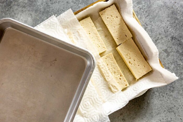 slices of tofu laid out on a paper towel-line baking sheet with another baking sheet set on top