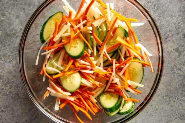 matchstick carrots, matchstick radish and thinly sliced cucumber in a large glass bowl with pickling liquid