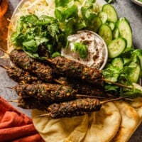 ground beef skewers on a platter with naan, cucumbers, shredded cabbage and a yogurt sauce