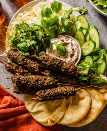 ground beef skewers on a platter with naan, cucumbers, shredded cabbage and a yogurt sauce