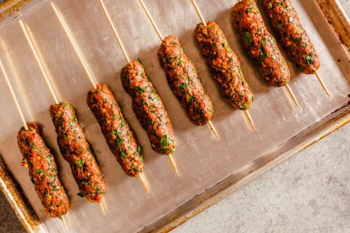 ground beef logs on wooden skewers on a baking sheet