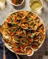 spaghetti with clams, breadcrumbs and diced red bell pepper and parsley in a large white bowl with serving spoons set in it and wine in short glasses set around