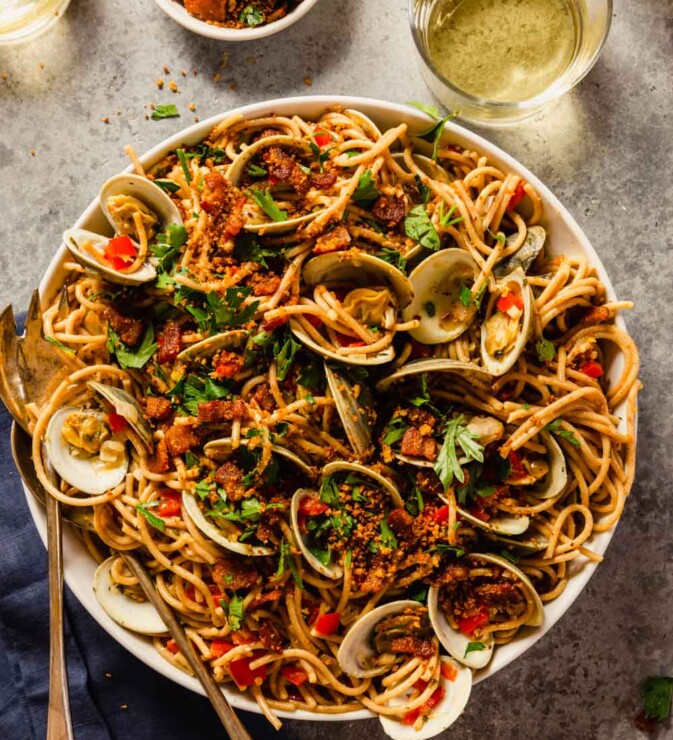 spaghetti with clams, breadcrumbs and diced red bell pepper and parsley in a large white bowl with serving spoons set in it and wine in short glasses set around