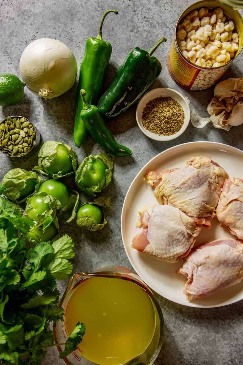 raw chicken thighs, tomatillos, cilantro, broth, chiles, hominy in a can, a white onion, a lime, garlic cloves, dried oregano and pumpkin seeds arranged on a counter
