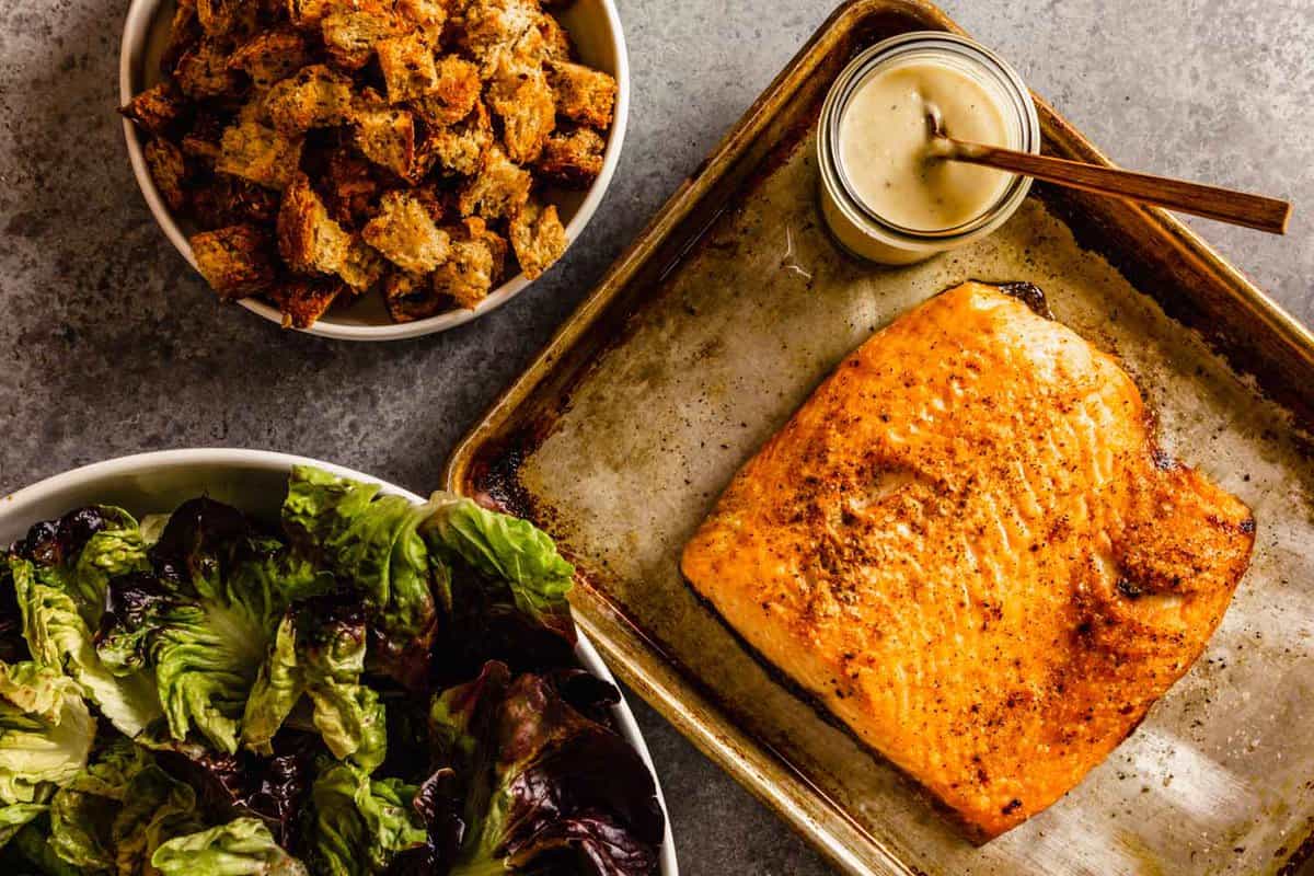 shallow white bowl filled with croutons set neat a baking sheet with broiled salmon and a bowl of greens