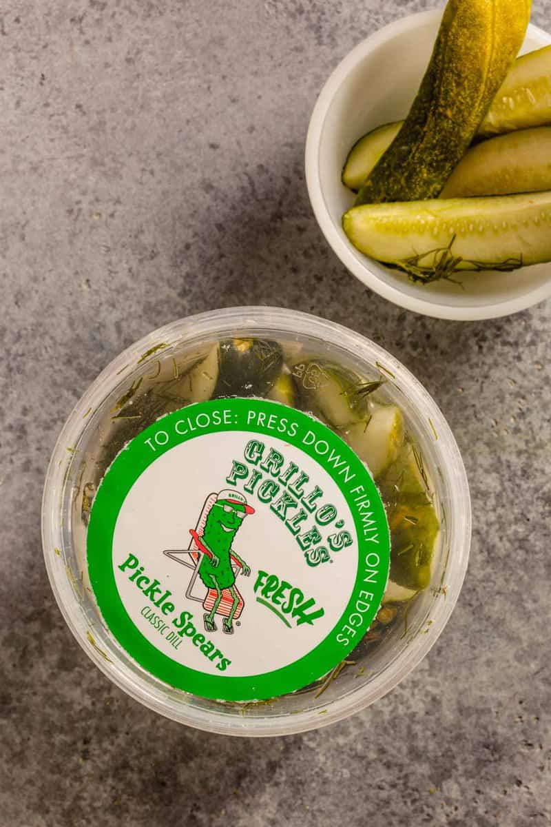 plastic container of dill pickle spears set on a counter with a small white bowl of pickle spears