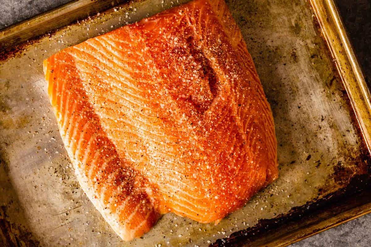 Large salmon fillet on a baking sheet with salt and pepper sprinkled over top.