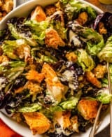 caesar salad with salmon and croutons in a large white bowl with two silver serving spoons