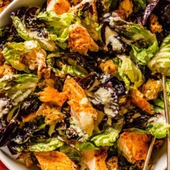 caesar salad with salmon and croutons in a large white bowl with two silver serving spoons