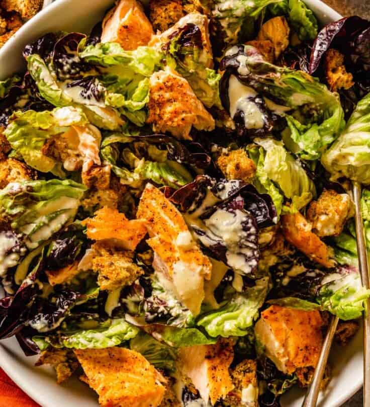 caesar salad with salmon and croutons in a large white bowl with two silver serving spoons