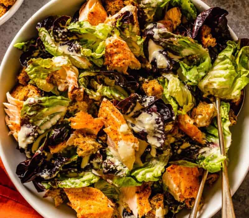 caesar salad with salmon and croutons in a large white bowl with two silver serving spoons
