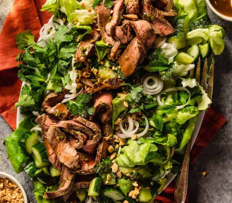 large white platter filled with lettuce greens and herbs, topped with slices of steak, thin slices of shallot, and chopped peanuts