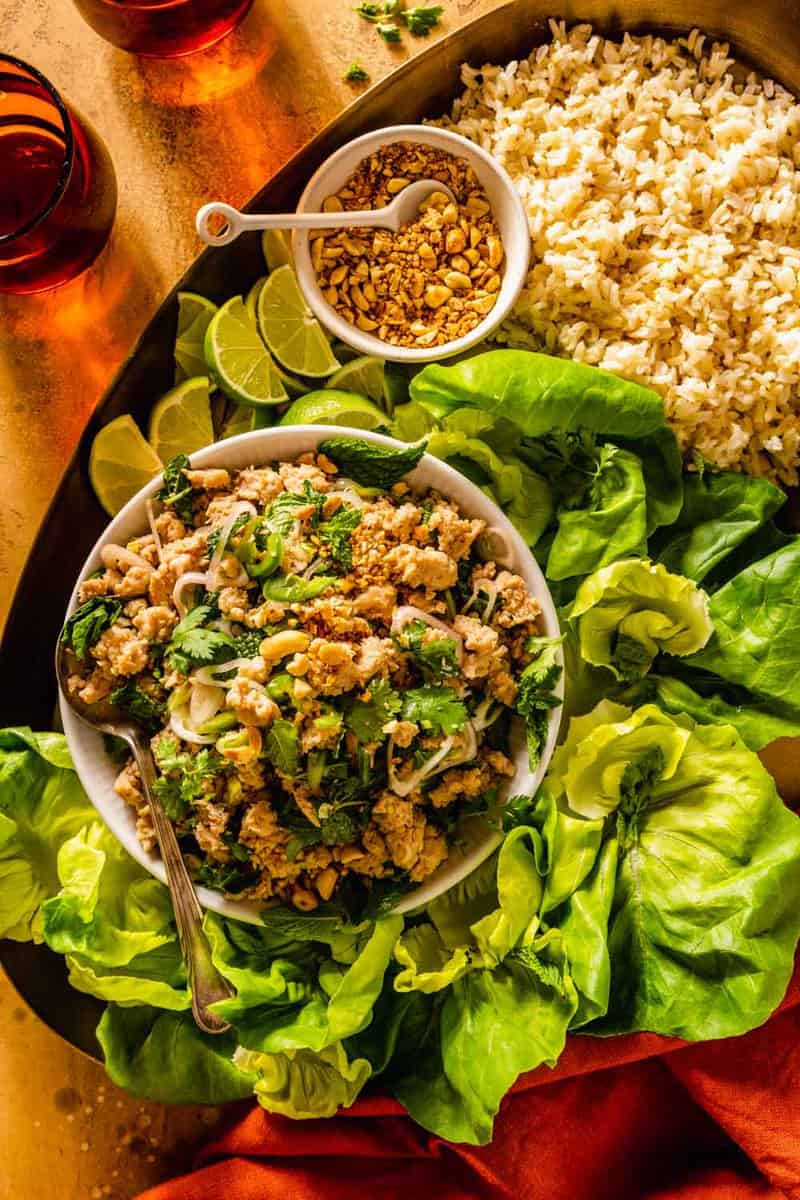 a shallow white bowl filled with ground chicken and herbs set on a platter with lettuce, rice, peanuts, and herbs