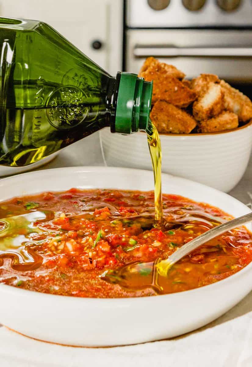olive oil getting drizzled over gazpacho in a shallow white bowl