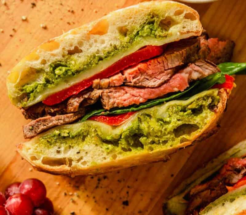 steak sandwich on a wood cutting board with pesto and grapes set around it