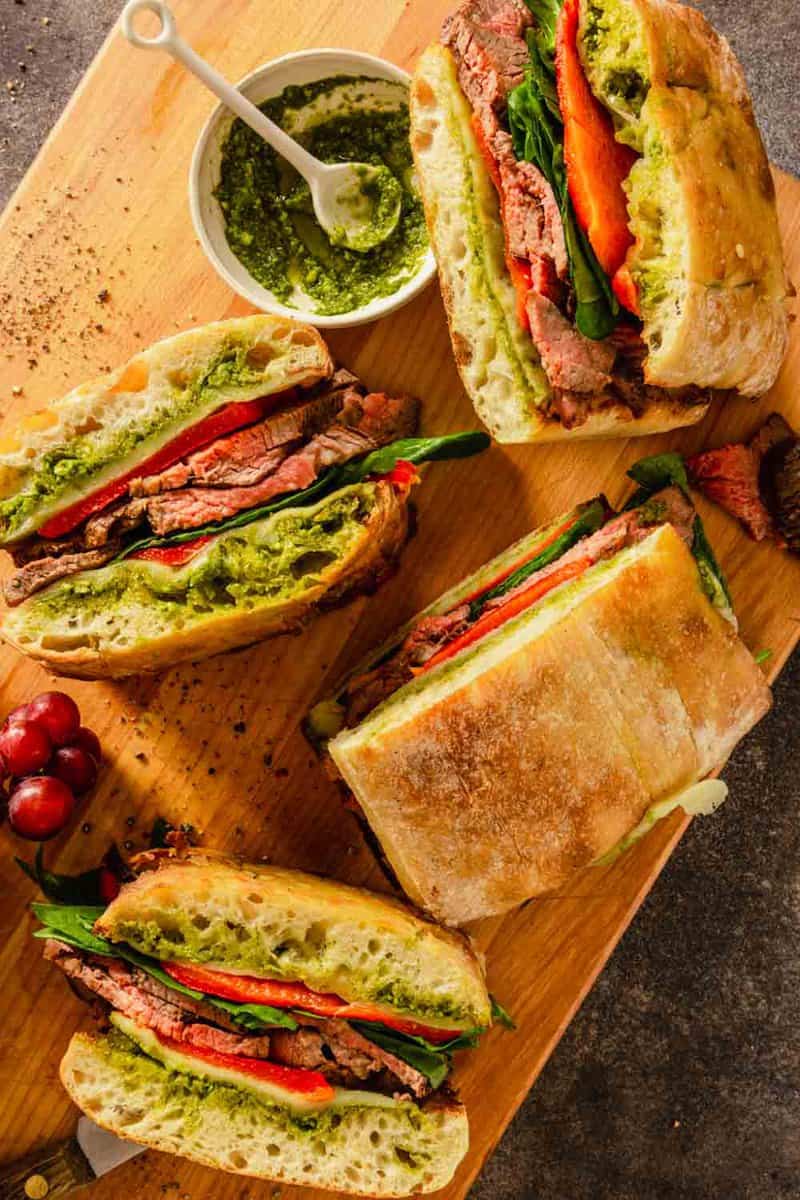 steak sandwiches on a wood cutting board with grapes and pesto set around them